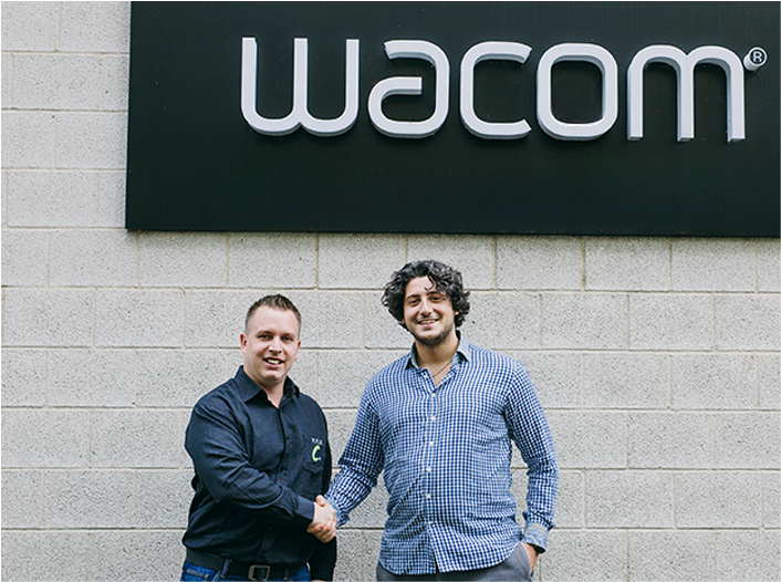 A picture of 2 men standing infront of a building with a Wacom sign, shaking hands